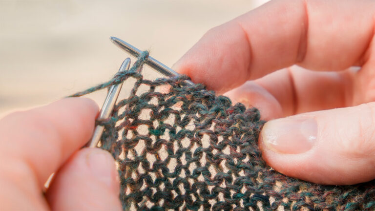 Friction is the key to cozy knits A close-up view of fingertips holding silver knitting needles, with light shining through the loose stitches in the cast fabric
