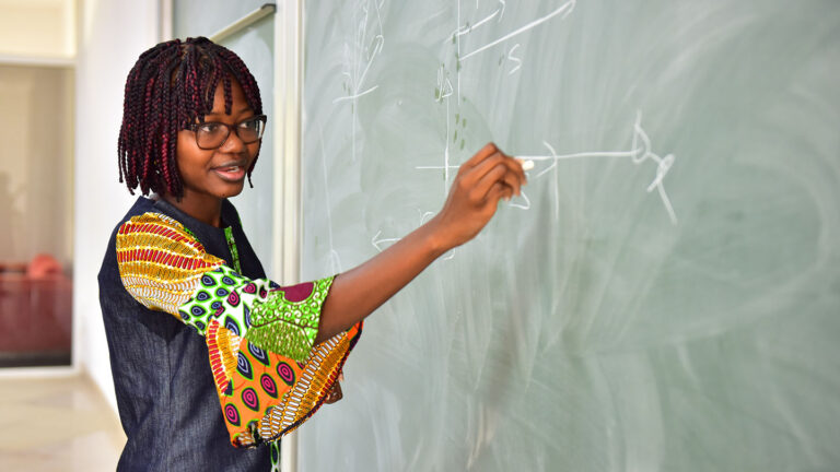 Meet one of the ‘world’s most interesting’ mathematicians Angela Tabiri at a green chalkboard. She has brown skin, her hair is styled in shoulder length braids and she wears a colorful dress. She also has glasses