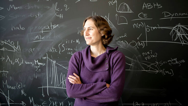 Cosmologist and dark matter researcher Katie Mack stands in front of a chalkboard covered in formulae. She is a white woman with short wavy brown hair and brown eyes.
